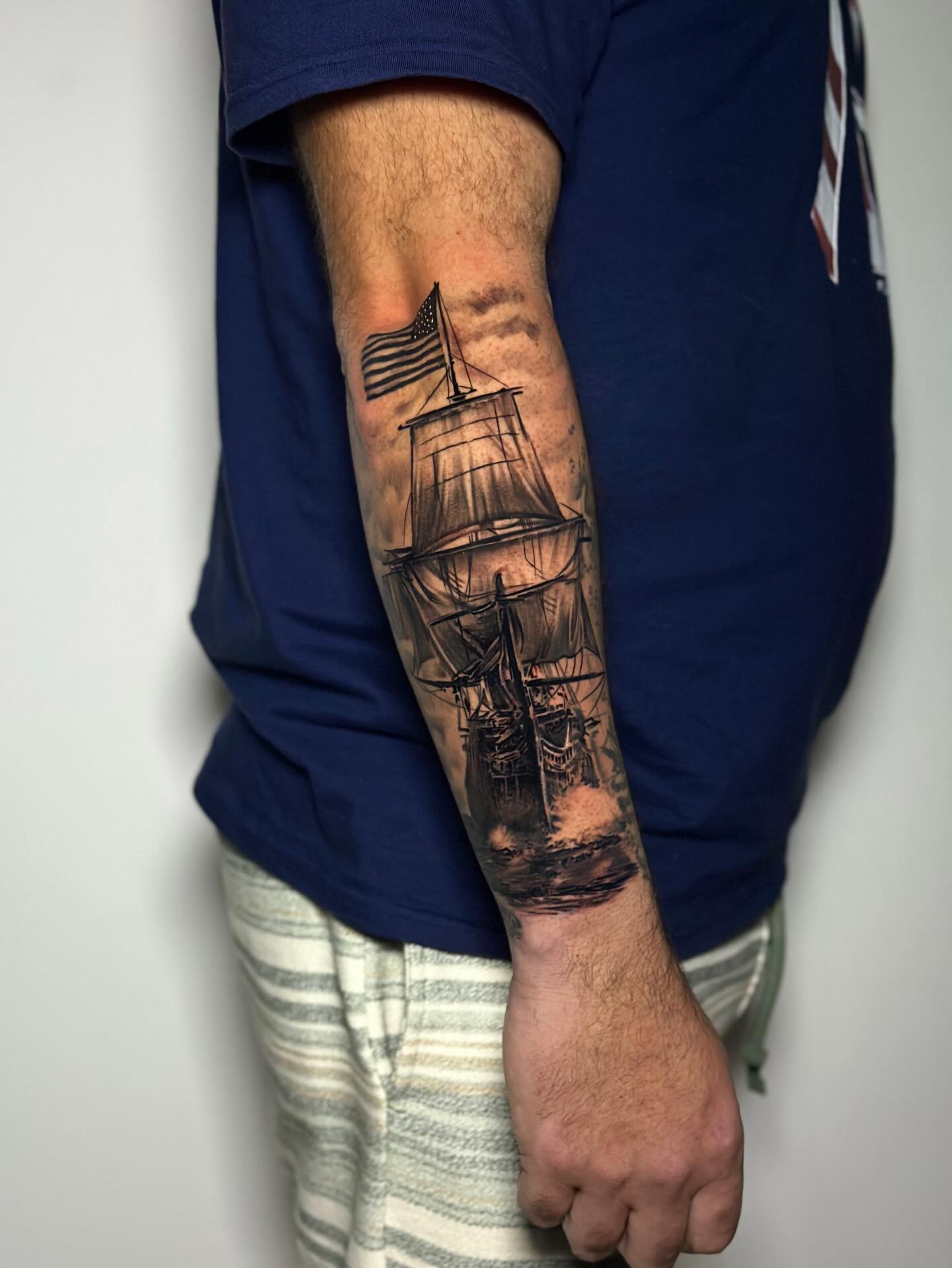 Tatuagem realista de navio à vela com bandeira americana no antebraço